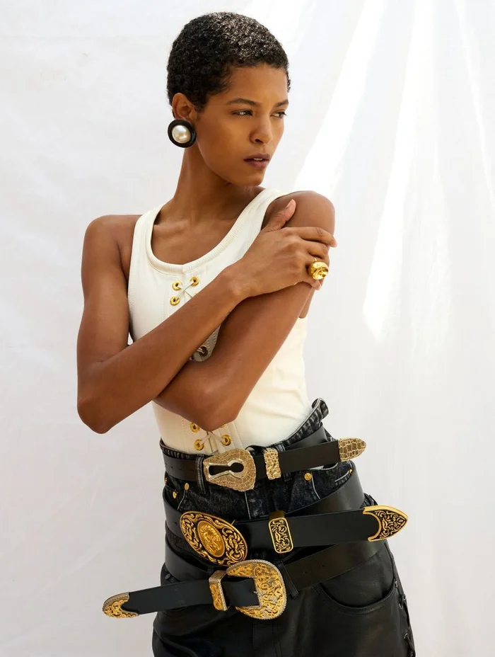 Model with short dark hair wearing a white tank top and multiple black leather belts with gold buckles against a white backdrop.