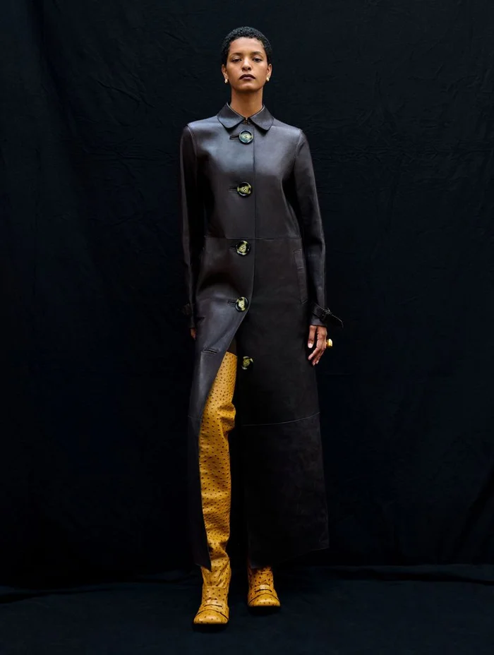 Model standing against dark fabric background wearing long dark leather coat and yellow boots