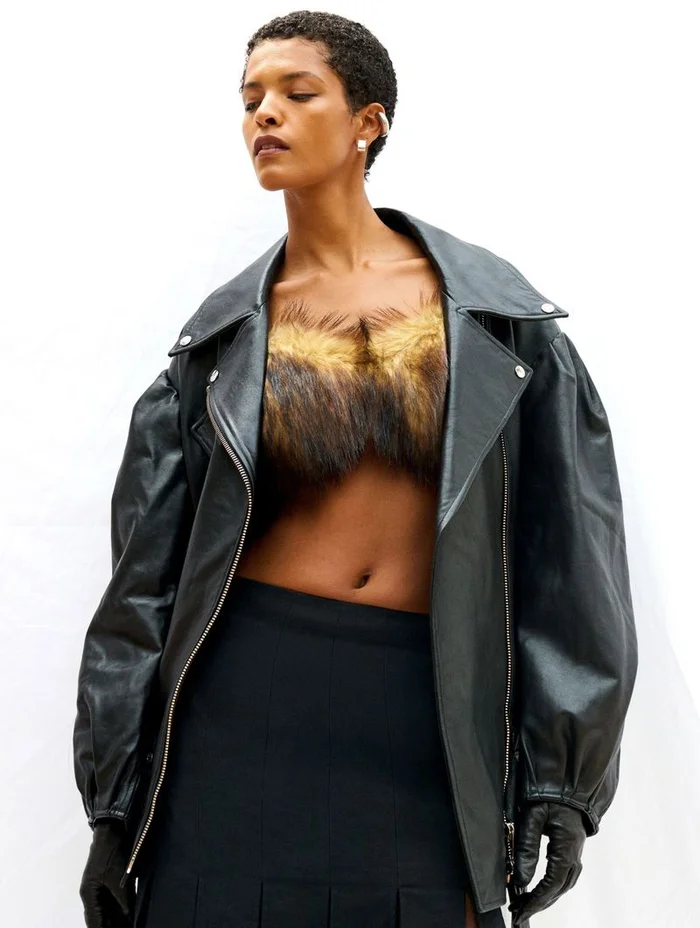 Model wearing a black leather jacket with faux fur accents and a black skirt against a plain white background