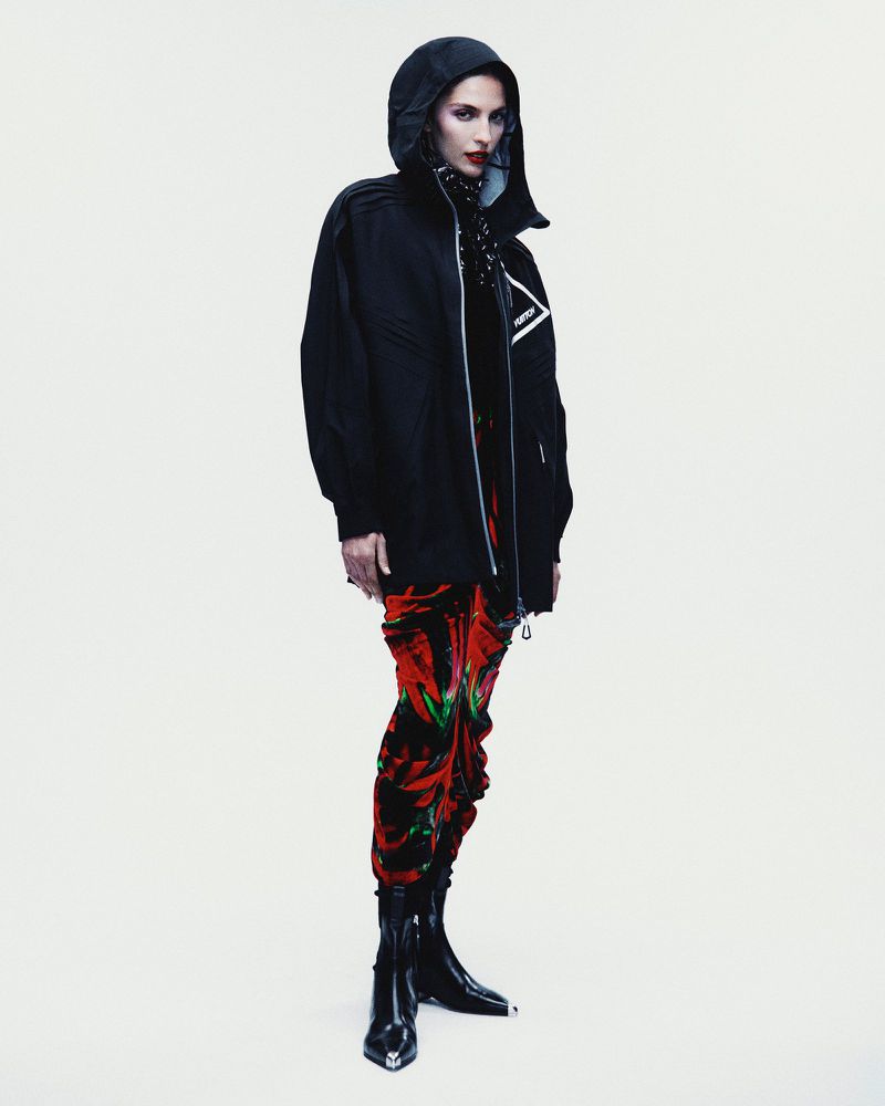 A woman in a black hooded jacket, patterned pants, and black boots strikes a full-body pose against a bright white backdrop.