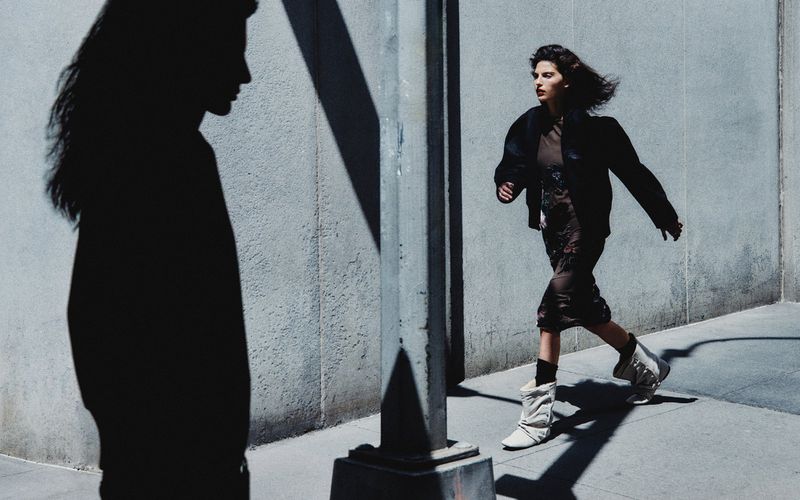 A woman in a dress and jacket is walking down a sidewalk in an urban setting with shadows, creating a dynamic and fashionable scene.