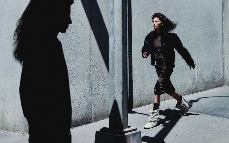 A woman in a dress and jacket is walking down a sidewalk in an urban setting with shadows, creating a dynamic and fashionable scene.