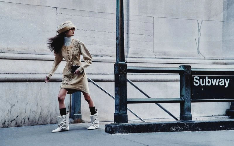 A woman in a vintage-style outfit walks near a subway entrance, featuring a tan dress, hat, white boots, and a city backdrop.