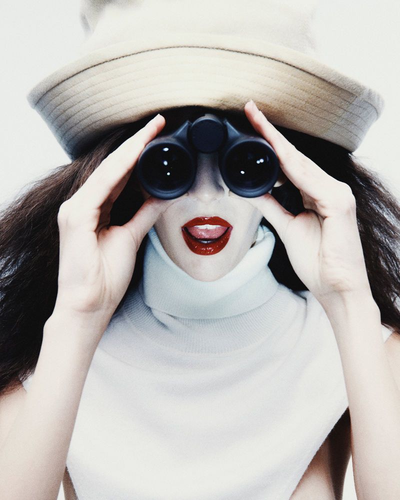 Woman in white turtleneck and hat looking through binoculars, standing against a white background with her mouth open slightly.