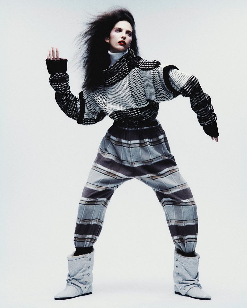 A fashion photograph presents a solitary adult female fashion model in a dynamic pose, showcasing a contemporary grey and black knitwear outfit.