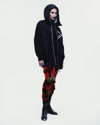 Model in a black hooded jacket and patterned trousers standing against a seamless white background for Vogue Mexico.