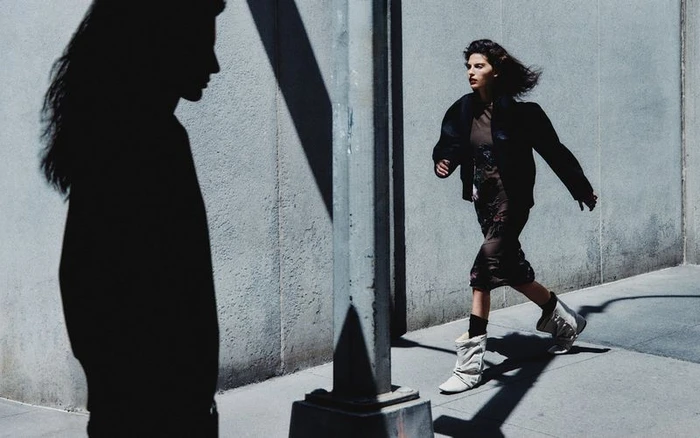 Model walking outdoors along a concrete wall with a silhouetted figure in the foreground