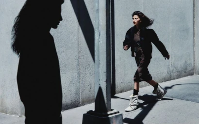 Model walking outdoors along a concrete wall with a silhouetted figure in the foreground
