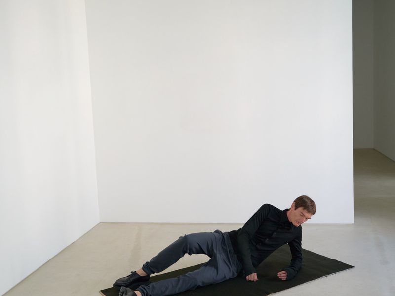 Adult male in athletic clothing lies on his side on a black mat, indoors, against a white wall, in a fitness or photo studio setting.