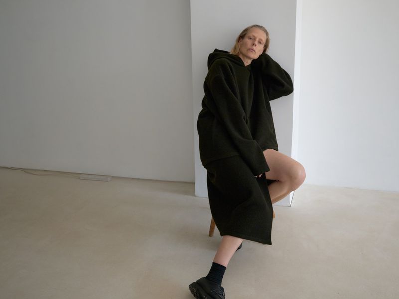Woman wearing a dark green oversized hoodie and skirt, sitting in a studio, with black socks and shoes.