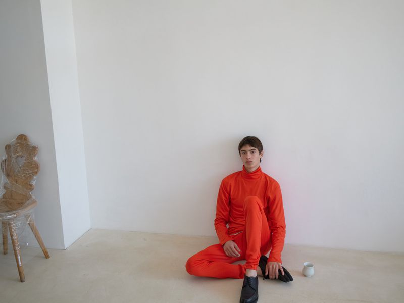 Young man sits on the floor in an orange outfit against a white wall, accompanied by a chair wrapped in plastic and a small white cup.