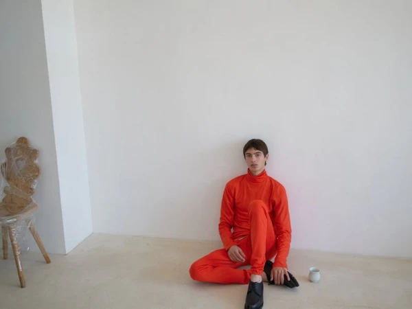 Young man sits on the floor in an orange outfit against a white wall, accompanied by a chair wrapped in plastic and a small white cup.
