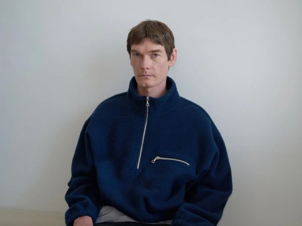 An adult male in a dark-blue fleece quarter-zip sweater in front of a plain white wall in a close-up studio portrait.