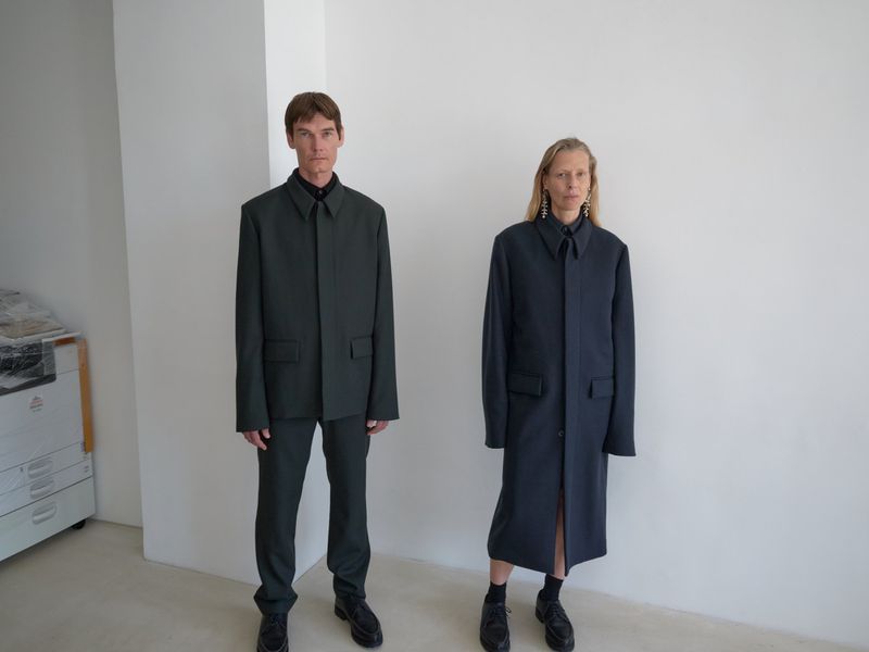 A man and woman stand in a minimalist white space, modeling fashion-forward outerwear. The studio lighting accentuates the garments.