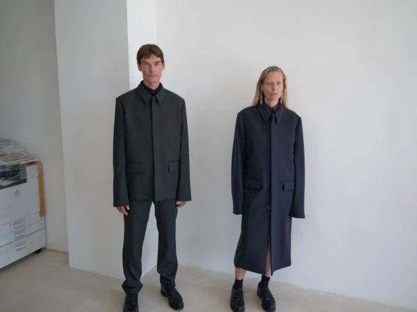 A man and woman stand in a minimalist white space, modeling fashion-forward outerwear. The studio lighting accentuates the garments.