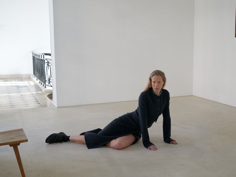 Woman in a black outfit poses on the floor in a minimalist white room, near a small wooden table and a window with black metal railings.