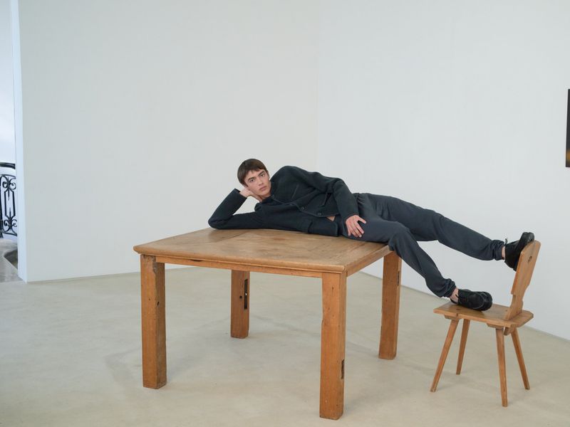 A brunette man in dark clothing reclines on a light wooden table with one foot resting on a chair in a minimalist white room.