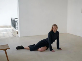 Model seated on a light floor wearing a dark long-sleeve top and matching skirt in a bright minimal interior