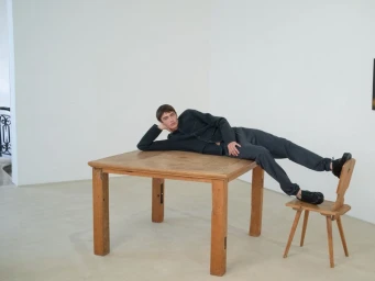 Young man lying on a large wooden table with his feet propped on a small chair in a bright gallery room