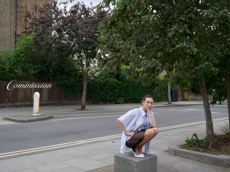 A woman squats on a concrete block in a street wearing a light blue oversized shirt, black skirt, and white heels under trees.