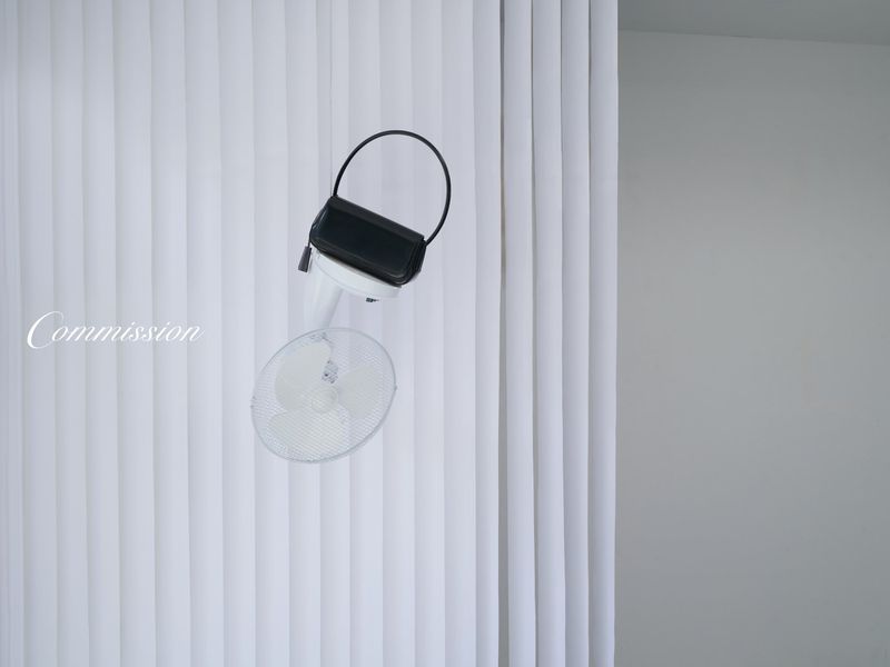 Fashion handbag on an electric fan against a minimalist backdrop featuring a white wall and flowing curtains in a studio setting.