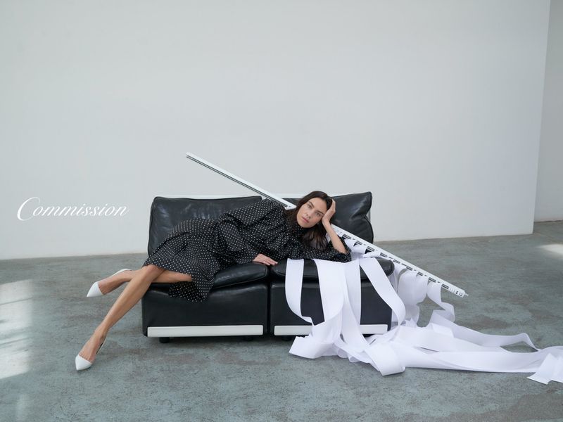 Adult woman lies on black couch with white high heels and wears a polka dot dress in a photo studio with white paper rolls.