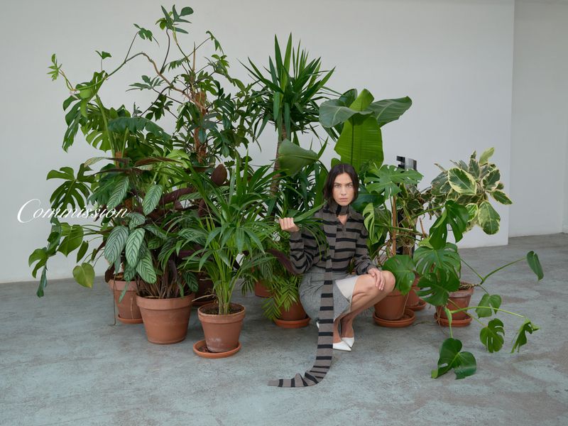 Woman poses kneeling among various potted plants in a studio, wearing a striped top, grey skirt, and white heels, with a light grey backdrop.