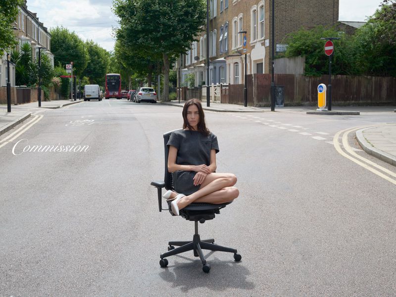 Woman in gray dress sits cross-legged on an office chair in the middle of a street with buildings and trees under a cloudy sky.