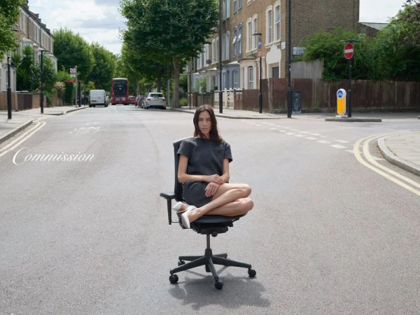 Woman in gray dress sits cross-legged on an office chair in the middle of a street with buildings and trees under a cloudy sky.