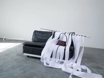 Dark leather sofa draped with long white ribbons and a burgundy handbag against a grey wall