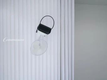 A black handbag resting on top of a white standing fan against a white vertically-ribbed studio backdrop.