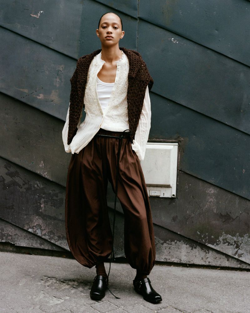 Fashionable adult woman wearing brown pants, white top and black shoes poses against a blue corrugated wall in natural daylight.
