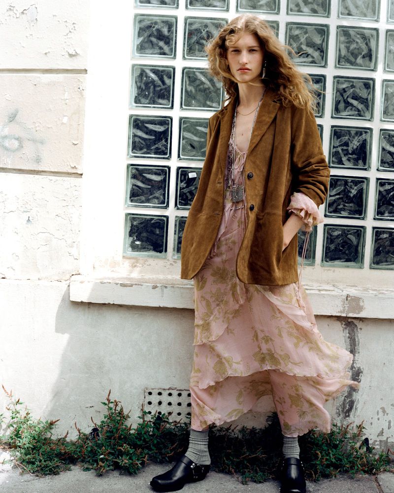A young woman with long brown hair wearing a brown blazer and floral print dress stands against a textured white wall outside.