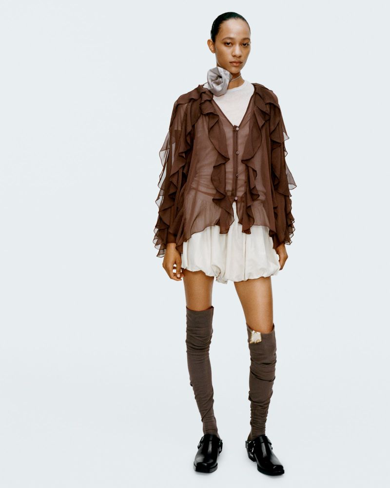 A slender woman poses in a sheer brown ruffled jacket, white shorts, brown thigh-high socks, and black Mary Jane shoes against a plain white background.