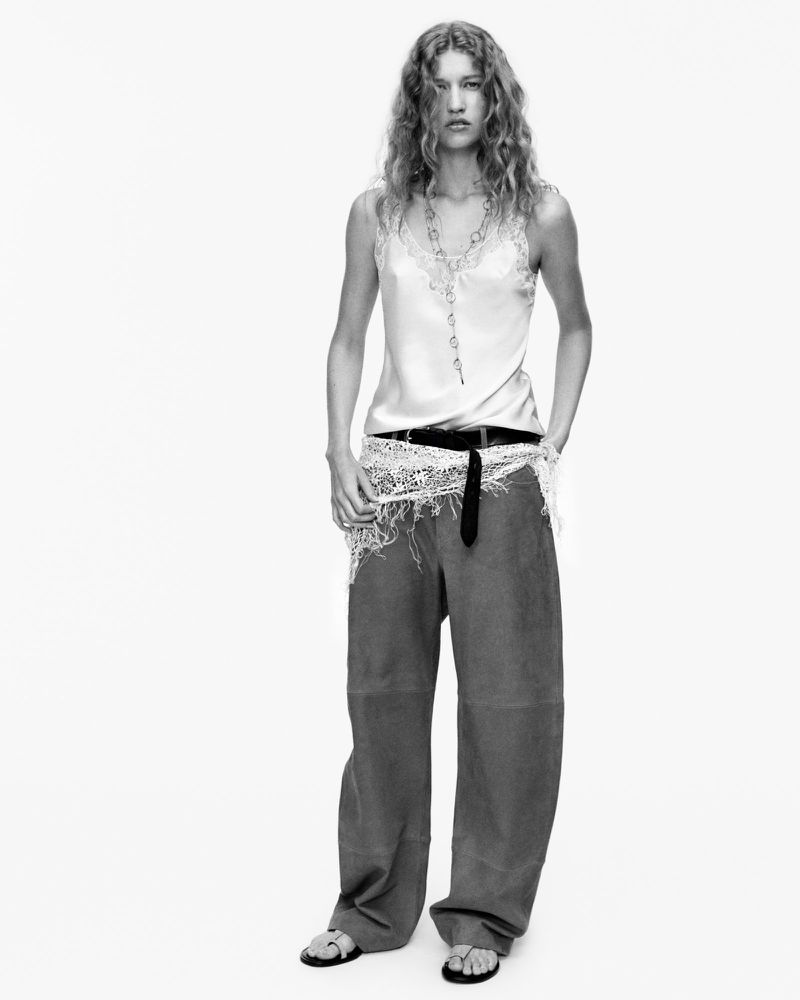 A model poses in a black and white fashion shot, wearing a tank top, loose trousers, and sandals against a stark white backdrop.