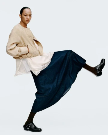 Fashion editorial shot of a model wearing a tan jacket, white top with lace trim, dark blue skirt, brown socks, and black shoes on white.