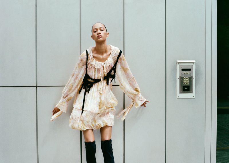 A woman in a patterned dress and black boots poses against a paneled wall with an intercom in a fashion photography shot.