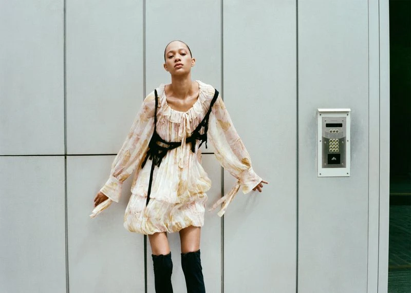 A woman in a patterned dress and black boots poses against a paneled wall with an intercom in a fashion photography shot.