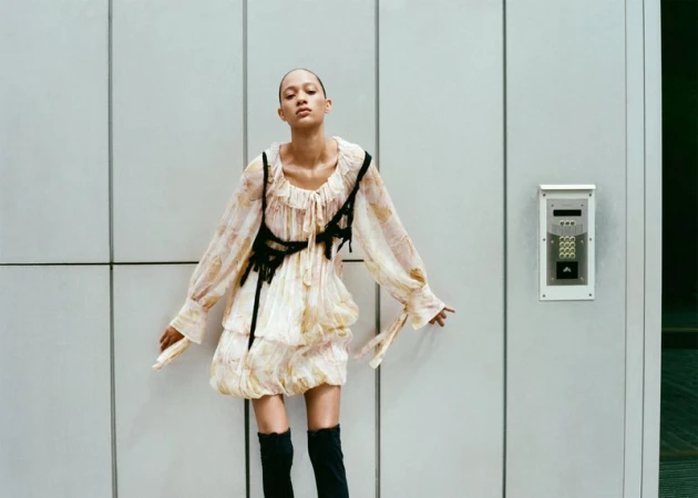A woman in a patterned dress and black boots poses against a paneled wall with an intercom in a fashion photography shot.