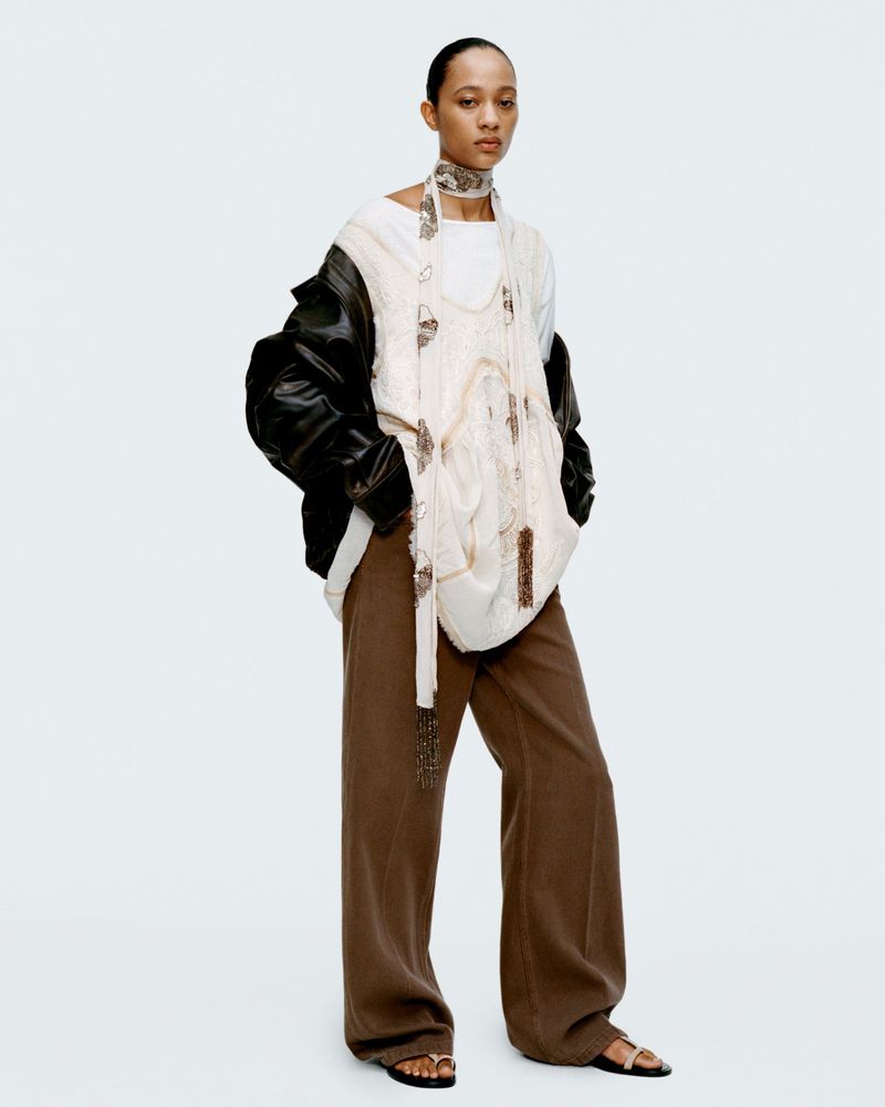Full shot of a woman in a white top, brown pants, and a black jacket, posing in a photo studio with a white backdrop.