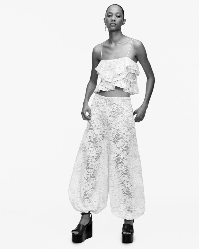 A Black female fashion model poses in a lacy white camisole and matching lace bloomers, along with black platform shoes, against a white backdrop.