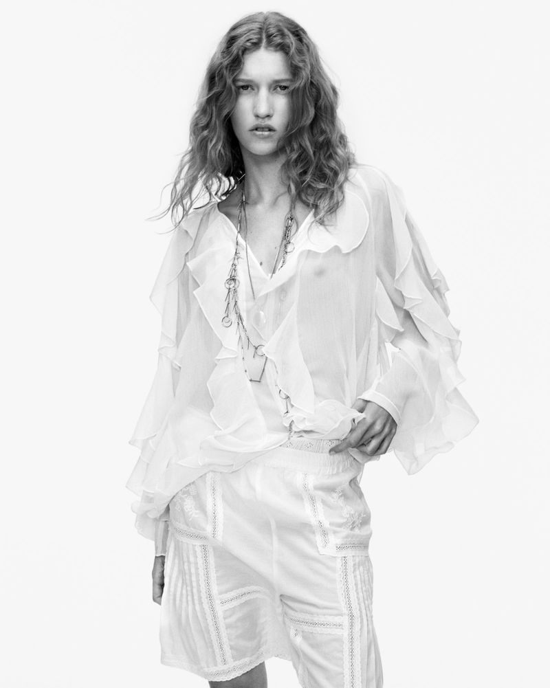 Black and white portrait of a model with wavy hair wearing a sheer ruffled blouse and shorts against a bright white background.