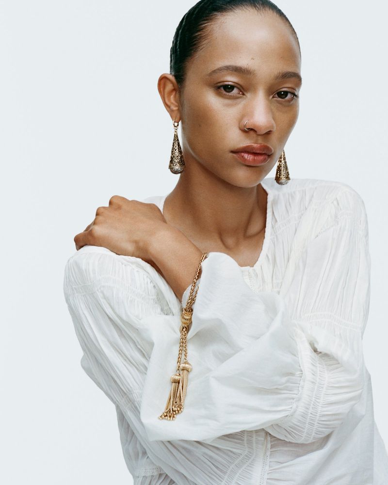 Close-up of a woman with medium skin tone wearing gold drop earrings and a white textured blouse, posing in a bright studio.