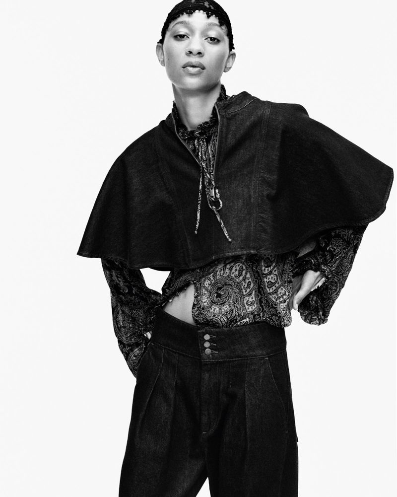 A black-and-white portrait of a model in a cape, paisley top, and wide-leg trousers, standing against a white backdrop in a studio shot.