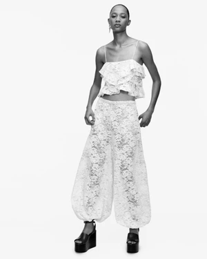 Model standing against a white backdrop wearing a white lace crop top and matching patterned trousers with platform wedge sandals.