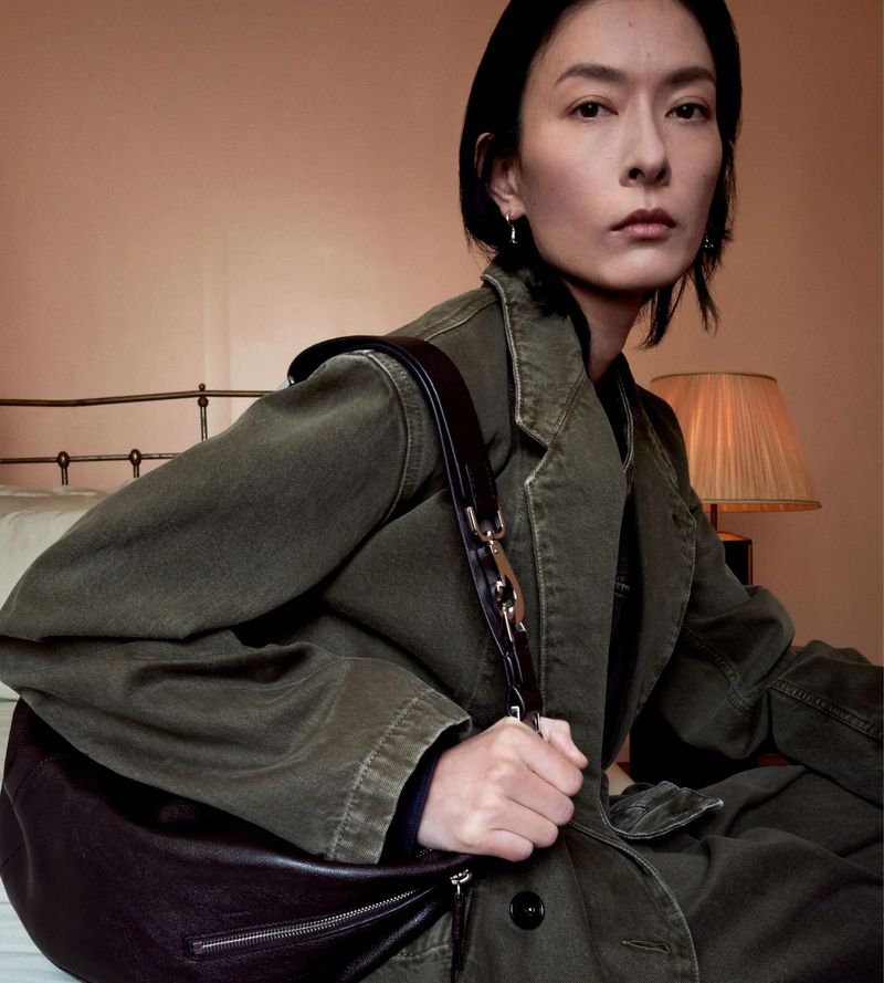 Asian female model showcasing a dark leather hobo bag in a casual indoor setting, wearing an olive green jacket on a bed with soft lighting.