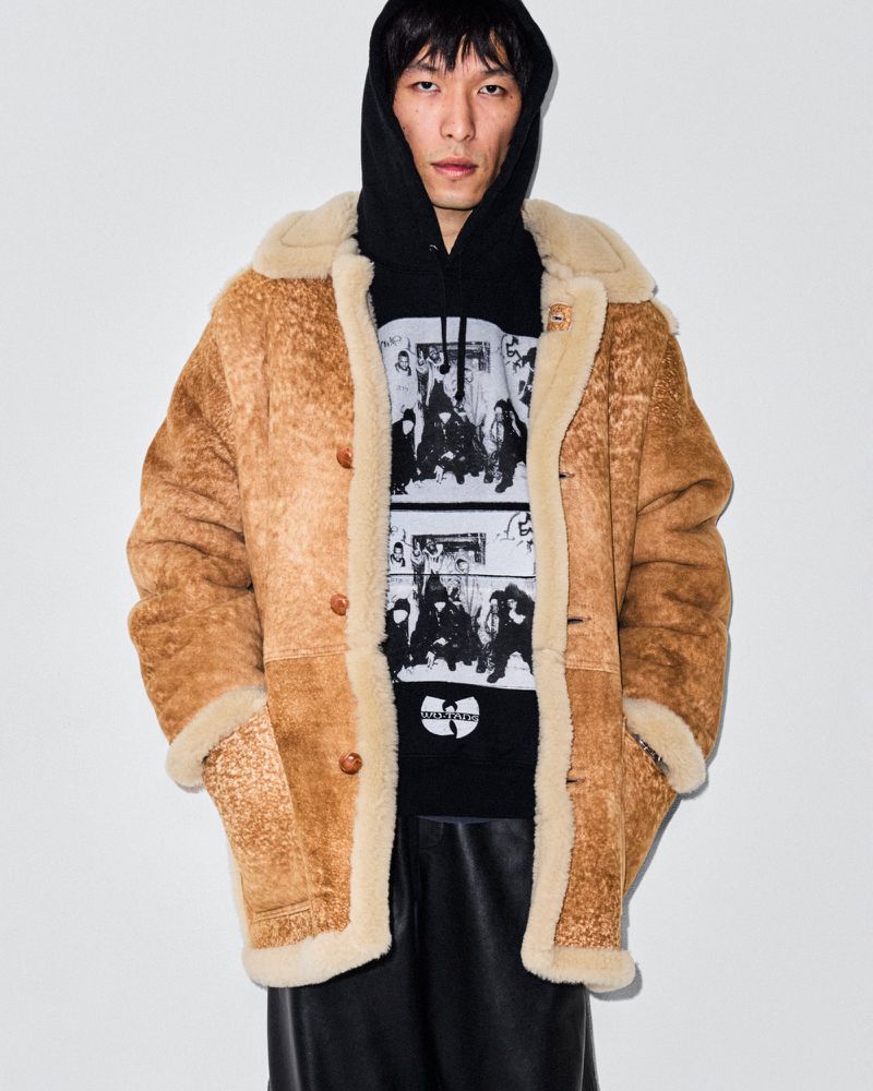 Man wearing a brown shearling coat and a black hoodie with a graphic print standing against a plain white backdrop.