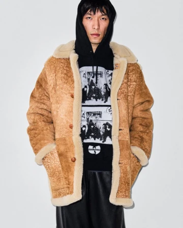 Man wearing a brown shearling coat and a black hoodie with a graphic print standing against a plain white backdrop.