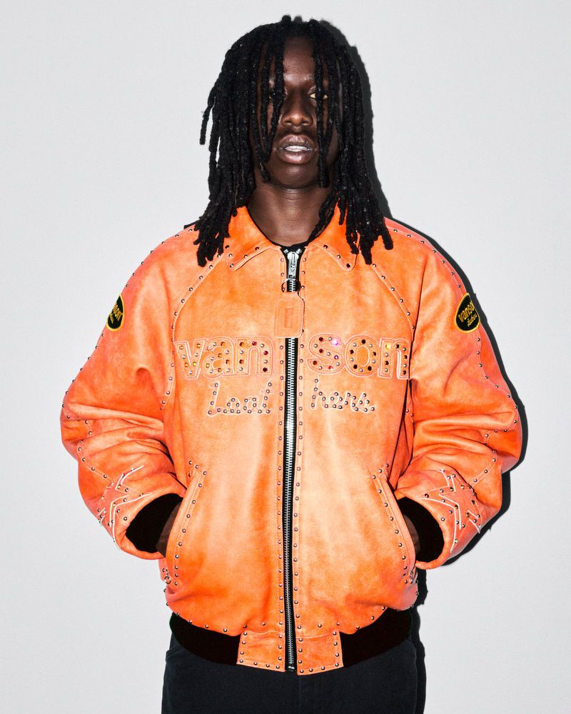 An adult Black male with dreadlocks poses indoors, wearing an orange leather bomber jacket with studded details against a white backdrop.