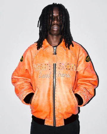 An adult Black male with dreadlocks poses indoors, wearing an orange leather bomber jacket with studded details against a white backdrop.